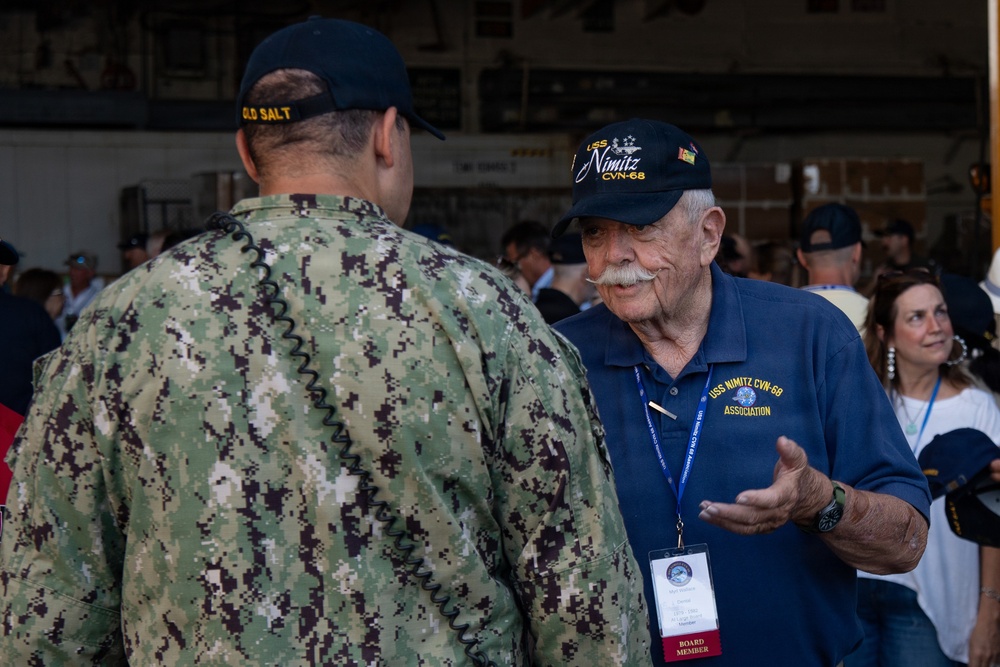 An elderly veteran wearing a USS Nimitz CVN-68 Association cap and polo shirt is engaged in conversation with an active-duty U.S. Navy sailor in NWU Type III camouflage uniform. The sailor's cap reads 'OLD SALT,' suggesting seniority or experience. The setting appears to be a naval facility or hangar bay during a reunion or commemorative event.