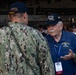 An elderly veteran wearing a USS Nimitz CVN-68 Association cap and polo shirt is engaged in conversation with an active-duty U.S. Navy sailor in NWU Type III camouflage uniform. The sailor's cap reads 'OLD SALT,' suggesting seniority or experience. The setting appears to be a naval facility or hangar bay during a reunion or commemorative event.