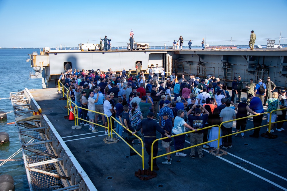A large crowd of civilians is gathered on the deck of a military naval vessel, likely during a public tour or open ship event. Several uniformed military personnel are visible on an upper deck level, with an American flag displayed. The event appears to be taking place at a harbor or naval base on a clear, sunny day.
