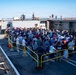 A large crowd of civilians is gathered on the deck of a military naval vessel, likely during a public tour or open ship event. Several uniformed military personnel are visible on an upper deck level, with an American flag displayed. The event appears to be taking place at a harbor or naval base on a clear, sunny day.