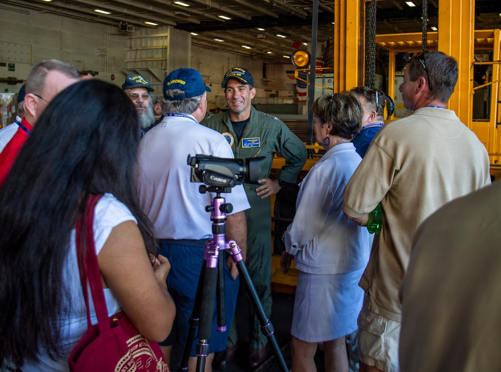 A military officer in a green flight suit is briefing or speaking to a group of civilian visitors inside what appears to be the hangar bay of a naval vessel. Several visitors are wearing navy-themed caps, and one person has set up a Canon camera on a tripod to document the event. The setting suggests an official tour or community outreach event aboard a U.S. Navy ship.