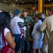 A military officer in a green flight suit is briefing or speaking to a group of civilian visitors inside what appears to be the hangar bay of a naval vessel. Several visitors are wearing navy-themed caps, and one person has set up a Canon camera on a tripod to document the event. The setting suggests an official tour or community outreach event aboard a U.S. Navy ship.
