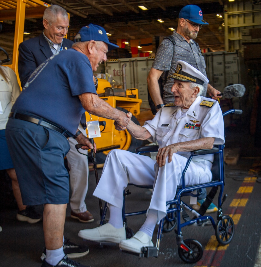 An elderly naval officer in full white dress uniform, seated in a wheelchair, is shaking hands with another older man wearing a blue t-shirt and cap. The setting appears to be the hangar deck of a naval vessel, with yellow equipment and shipping containers visible in the background. Several other individuals are present, including a man in a suit and another wearing a Chicago Cubs cap.