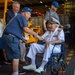 An elderly naval officer in full white dress uniform, seated in a wheelchair, is shaking hands with another older man wearing a blue t-shirt and cap. The setting appears to be the hangar deck of a naval vessel, with yellow equipment and shipping containers visible in the background. Several other individuals are present, including a man in a suit and another wearing a Chicago Cubs cap.