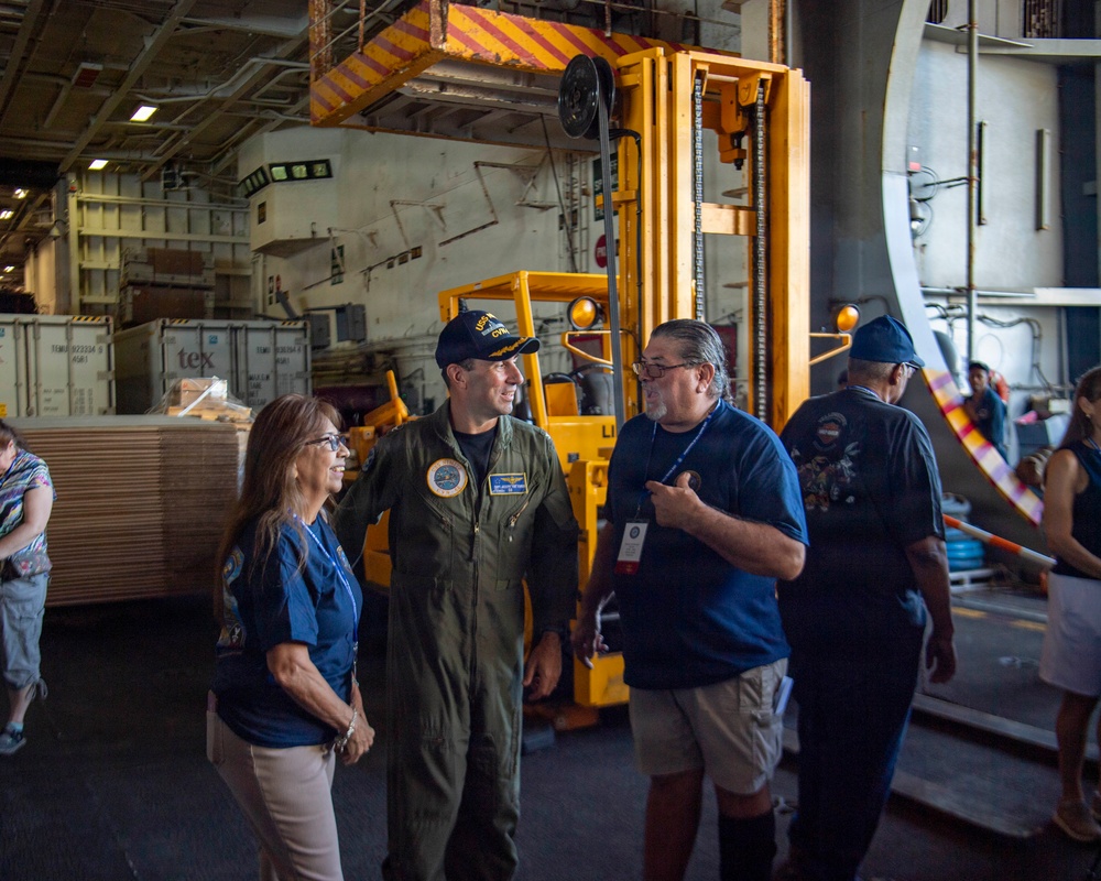 A military officer in a green flight suit and navy cap is engaged in conversation with two civilians wearing matching blue shirts inside what appears to be the hangar bay of a naval vessel. The setting shows industrial equipment including a yellow forklift and shipping containers in the background. This appears to be a tour or official visit event, as indicated by the visitor badge worn by one of the civilians.