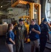 A military officer in a green flight suit and navy cap is engaged in conversation with two civilians wearing matching blue shirts inside what appears to be the hangar bay of a naval vessel. The setting shows industrial equipment including a yellow forklift and shipping containers in the background. This appears to be a tour or official visit event, as indicated by the visitor badge worn by one of the civilians.