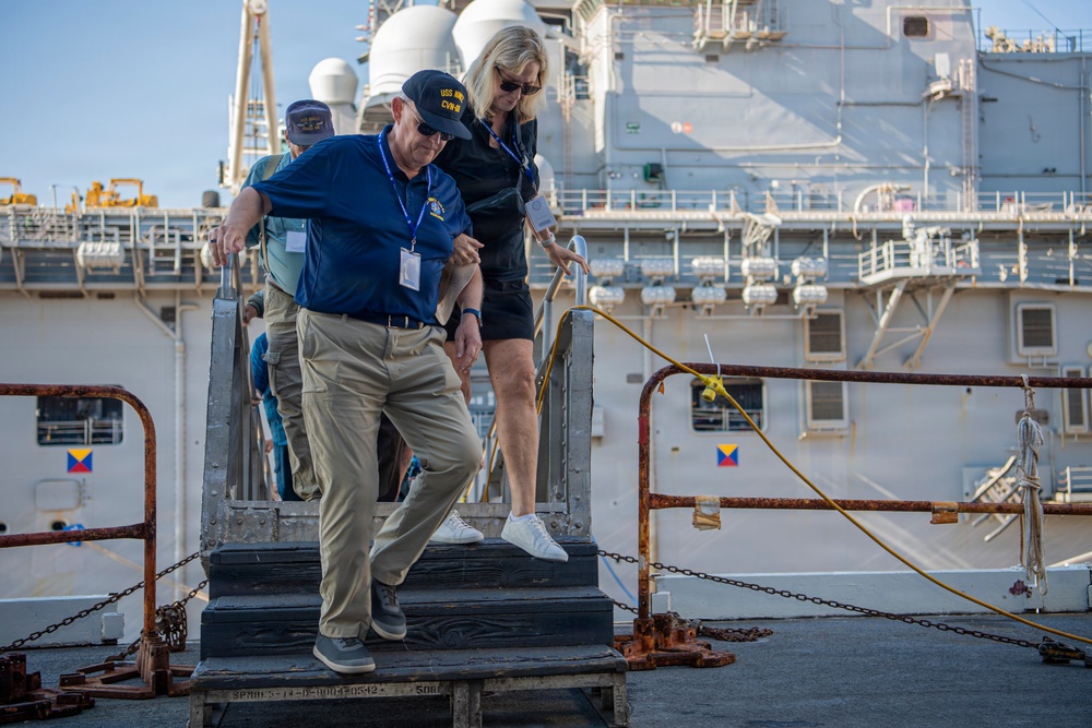 Two civilians are descending metal steps on what appears to be a naval pier or dock area, with a large warship visible in the background. The man in the foreground is wearing a blue polo shirt, khaki pants, and a USS Abraham Lincoln (CVN-3) cap, along with a blue lanyard/badge indicating a visitor or official pass. The setting appears to be an official naval facility visit or tour.