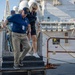 Two civilians are descending metal steps on what appears to be a naval pier or dock area, with a large warship visible in the background. The man in the foreground is wearing a blue polo shirt, khaki pants, and a USS Abraham Lincoln (CVN-3) cap, along with a blue lanyard/badge indicating a visitor or official pass. The setting appears to be an official naval facility visit or tour.