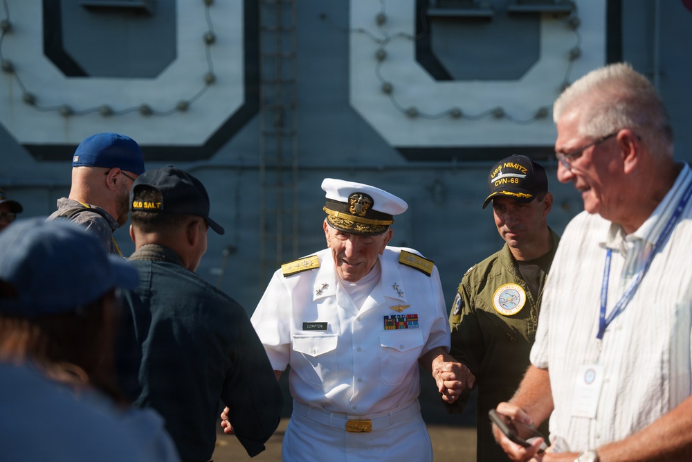 A senior U.S. Navy officer in formal white dress uniform, identified by name tag as 'Compton', is engaged in conversation with a group of individuals near what appears to be a naval vessel. The gathering includes personnel in various uniforms and civilians, with one person wearing a USS Nimitz CVN-68 cap visible in the background. The setting appears to be an outdoor naval event or ceremony.