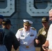 A senior U.S. Navy officer in formal white dress uniform, identified by name tag as 'Compton', is engaged in conversation with a group of individuals near what appears to be a naval vessel. The gathering includes personnel in various uniforms and civilians, with one person wearing a USS Nimitz CVN-68 cap visible in the background. The setting appears to be an outdoor naval event or ceremony.