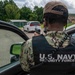 A U.S. Navy Security Forces personnel is shown from behind, wearing a black tactical vest labeled 'U.S. NAVY SECURITY FORCES' over digital camouflage uniform. The individual is holding a radio/communication device and appears to be conducting a vehicle stop or checkpoint inspection of a white minivan in a parking area. The setting appears to be a military installation based on the surrounding buildings and infrastructure.
