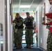 The image shows U.S. Navy personnel conducting what appears to be a tactical training exercise or drill inside a building corridor. Two armed personnel in Navy camouflage uniforms and tactical vests are advancing down a hallway with weapons raised, while a third person wearing a red safety vest and holding a clipboard observes and evaluates the exercise. Yellow caution tape and safety cones are visible, indicating a controlled training environment.
