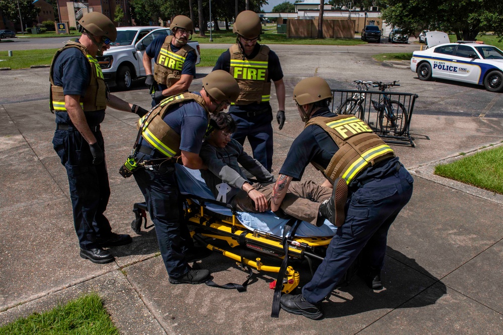 A group of firefighters wearing tactical gear, ballistic helmets, and body armor are loading or attending to a patient on a yellow Stryker stretcher in an outdoor parking lot. The scene appears to be a tactical emergency medical response drill or training exercise, indicated by the controlled environment and U.S. Navy Police vehicle visible in the background. Multiple fire personnel in navy blue uniforms with yellow reflective striping are actively engaged with the patient.