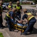 A group of firefighters wearing tactical gear, ballistic helmets, and body armor are loading or attending to a patient on a yellow Stryker stretcher in an outdoor parking lot. The scene appears to be a tactical emergency medical response drill or training exercise, indicated by the controlled environment and U.S. Navy Police vehicle visible in the background. Multiple fire personnel in navy blue uniforms with yellow reflective striping are actively engaged with the patient.