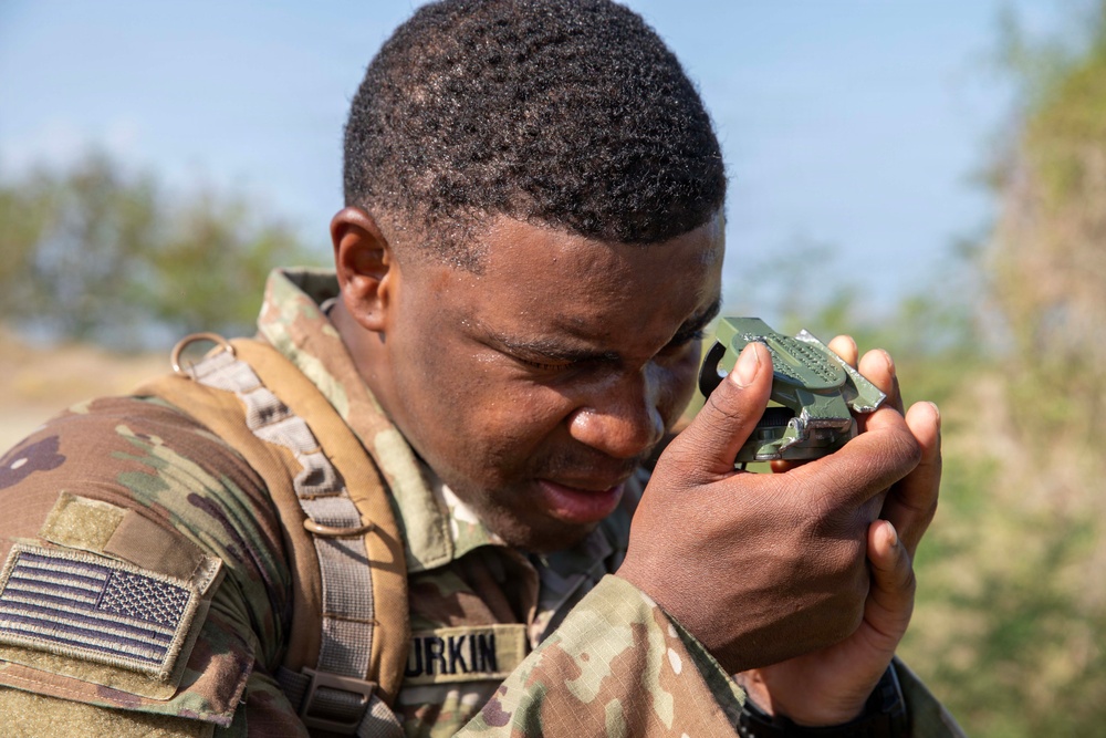 A U.S. Army soldier in MultiCam (OCP) camouflage uniform is closely examining a lensatic compass, likely taking a bearing or azimuth reading during a land navigation exercise. The soldier's name tape reads 'URKIN' (partially visible) and an American flag patch is visible on the left shoulder. The image appears to be taken outdoors in a training environment.