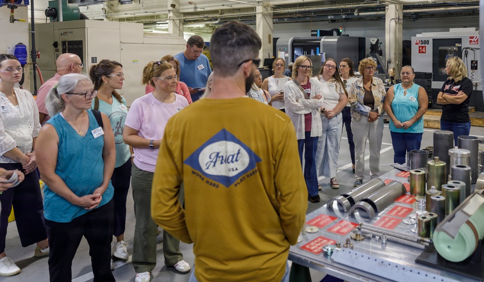 A group of approximately 20 adults are gathered in an industrial manufacturing facility, listening to a presenter wearing a mustard-yellow Ariat brand long-sleeve shirt. The setting appears to be a factory tour or educational visit, with CNC machining equipment visible in the background. Several participants are wearing name badges, suggesting a formal organized event.