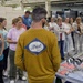 A group of approximately 20 adults are gathered in an industrial manufacturing facility, listening to a presenter wearing a mustard-yellow Ariat brand long-sleeve shirt. The setting appears to be a factory tour or educational visit, with CNC machining equipment visible in the background. Several participants are wearing name badges, suggesting a formal organized event.