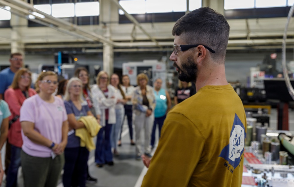 A man in a yellow shirt wearing safety glasses appears to be giving a tour or presentation to a group of people in an industrial or manufacturing facility. The group, consisting mostly of women, is listening attentively while standing in front of industrial machinery. The setting appears to be a factory floor or machine shop with CNC equipment visible in the background.