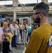 A man in a yellow shirt wearing safety glasses appears to be giving a tour or presentation to a group of people in an industrial or manufacturing facility. The group, consisting mostly of women, is listening attentively while standing in front of industrial machinery. The setting appears to be a factory floor or machine shop with CNC equipment visible in the background.