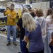A male worker in a yellow long-sleeve shirt and safety glasses is presenting or demonstrating something to a group of visitors inside an industrial manufacturing facility. He appears to be holding a small object while addressing approximately 8-10 people, predominantly women, who are attentively listening. The setting contains CNC machines, including visible Okuma brand equipment, suggesting a machining or manufacturing plant tour.