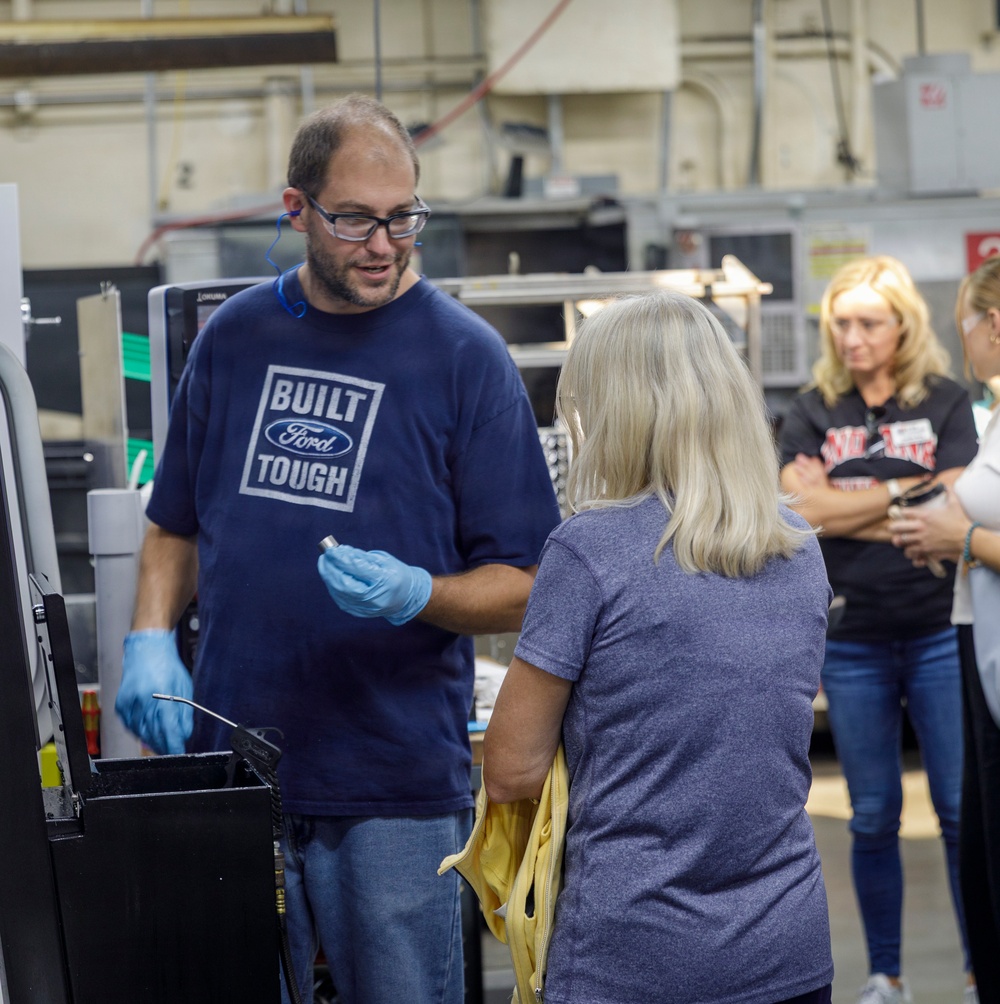A man wearing a 'Built Ford Tough' navy t-shirt and blue latex gloves appears to be demonstrating or explaining something to a small group of visitors in an industrial or manufacturing facility. He is holding a small metal component and speaking to a woman with gray-blonde hair, while two other women observe in the background. The setting appears to be a factory or machine shop tour.