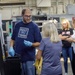 A man wearing a 'Built Ford Tough' navy t-shirt and blue latex gloves appears to be demonstrating or explaining something to a small group of visitors in an industrial or manufacturing facility. He is holding a small metal component and speaking to a woman with gray-blonde hair, while two other women observe in the background. The setting appears to be a factory or machine shop tour.
