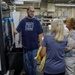 A male worker wearing a 'Built Ford Tough' t-shirt and blue nitrile gloves is demonstrating or explaining an Okuma CNC machine to a small group of visitors in an industrial manufacturing facility. The setting appears to be a factory tour or educational visit, with several women listening attentively to the worker's explanation. The environment is a well-lit manufacturing floor with various industrial equipment visible in the background.