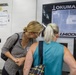 Two women are examining an Okuma LU4000 EX SimulTurn CNC lathe machine in an industrial manufacturing facility. One woman wearing a checkered shirt and safety glasses appears to be explaining or pointing something out, while the other in a teal sleeveless top observes the machine. The setting suggests a factory tour or training session.