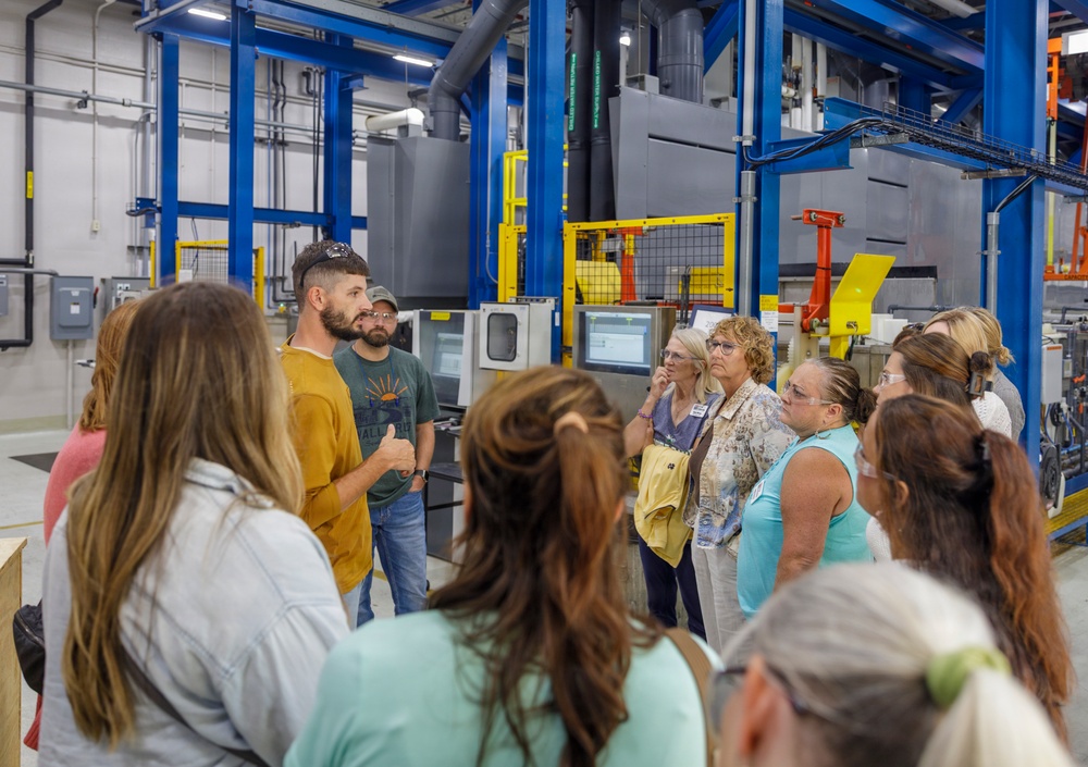 A group of approximately 12-15 adults are participating in a guided facility tour inside an industrial manufacturing or processing plant. A man in a mustard-yellow shirt appears to be the tour guide, speaking and gesturing to the group. The facility features large blue steel structural frames, gray industrial machinery, yellow safety barriers, and overhead ductwork.