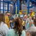 A group of approximately 12-15 adults are participating in a guided facility tour inside an industrial manufacturing or processing plant. A man in a mustard-yellow shirt appears to be the tour guide, speaking and gesturing to the group. The facility features large blue steel structural frames, gray industrial machinery, yellow safety barriers, and overhead ductwork.