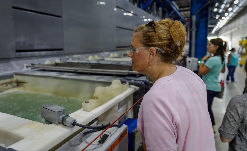 A woman wearing safety glasses and a pink sweatshirt is closely observing an industrial processing tank or bath in what appears to be a manufacturing facility. Several other people are visible in the background, suggesting this may be a tour or inspection visit. The facility features large industrial equipment, blue structural supports, and overhead lighting.