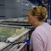 A woman wearing safety glasses and a pink sweatshirt is closely observing an industrial processing tank or bath in what appears to be a manufacturing facility. Several other people are visible in the background, suggesting this may be a tour or inspection visit. The facility features large industrial equipment, blue structural supports, and overhead lighting.