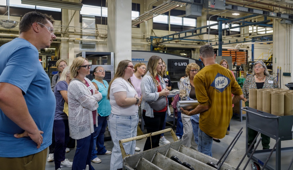 A group of approximately 12-15 people are gathered in an industrial manufacturing facility, appearing to be on a guided tour or informational presentation. A man in a mustard-yellow shirt with a 'Fiat' logo is facing the group, seemingly explaining or demonstrating something near industrial equipment. The setting includes visible sheet metal fabrication machinery, including an LVD STRIPPIT punch/laser machine.