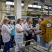 A group of approximately 12-15 people are gathered in an industrial manufacturing facility, appearing to be on a guided tour or informational presentation. A man in a mustard-yellow shirt with a 'Fiat' logo is facing the group, seemingly explaining or demonstrating something near industrial equipment. The setting includes visible sheet metal fabrication machinery, including an LVD STRIPPIT punch/laser machine.