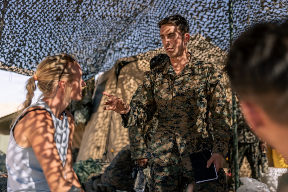 A military personnel wearing MARPAT (Marine pattern) camouflage uniform is engaged in conversation with a civilian woman with blonde hair in a ponytail. The interaction appears to take place under a camouflage netting structure, suggesting a field operations or forward operating base setting. A third partially visible individual is present in the foreground right.