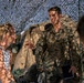A military personnel wearing MARPAT (Marine pattern) camouflage uniform is engaged in conversation with a civilian woman with blonde hair in a ponytail. The interaction appears to take place under a camouflage netting structure, suggesting a field operations or forward operating base setting. A third partially visible individual is present in the foreground right.