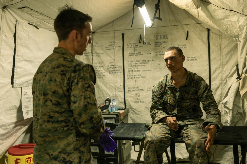 Two military personnel in MARPAT camouflage uniforms are inside a field medical tent. One individual is seated on what appears to be an examination table while the other stands facing them, suggesting a medical consultation or assessment. The tent walls display handwritten medical reference charts, including one clearly labeled 'HEAT INJURIES'.