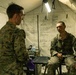 Two military personnel in MARPAT camouflage uniforms are inside a field medical tent. One individual is seated on what appears to be an examination table while the other stands facing them, suggesting a medical consultation or assessment. The tent walls display handwritten medical reference charts, including one clearly labeled 'HEAT INJURIES'.