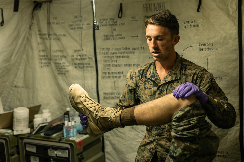 A U.S. Marine in MARPAT camouflage uniform wearing purple nitrile medical gloves is examining a boot on a patient's foot inside a military medical tent. The background displays handwritten medical reference charts, including one titled 'HEAT INJURIES,' indicating this is a field medical training or treatment environment. Medical supplies and equipment are visible on a table to the left.