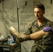 A U.S. Marine in MARPAT camouflage uniform wearing purple nitrile medical gloves is examining a boot on a patient's foot inside a military medical tent. The background displays handwritten medical reference charts, including one titled 'HEAT INJURIES,' indicating this is a field medical training or treatment environment. Medical supplies and equipment are visible on a table to the left.