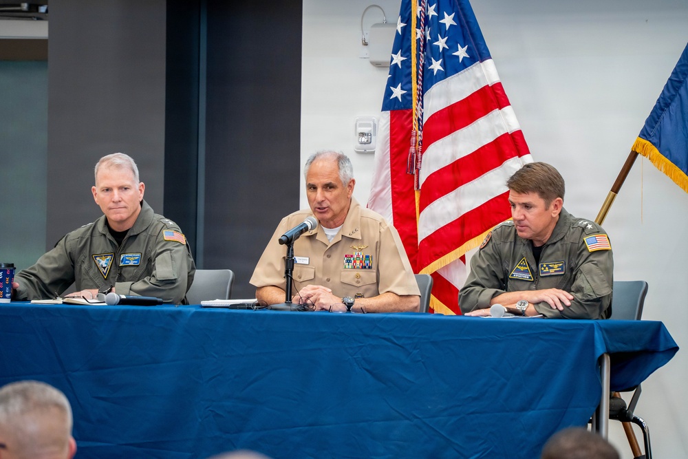 Three U.S. military officers are seated at a blue-draped table during what appears to be a formal briefing or press conference. The center officer, wearing a tan Navy uniform with multiple service ribbons, is speaking into a microphone. The flanking officers wear olive/sage green flight suits with unit patches and U.S. flag patches.