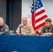Three U.S. military officers are seated at a blue-draped table during what appears to be a formal briefing or press conference. The center officer, wearing a tan Navy uniform with multiple service ribbons, is speaking into a microphone. The flanking officers wear olive/sage green flight suits with unit patches and U.S. flag patches.