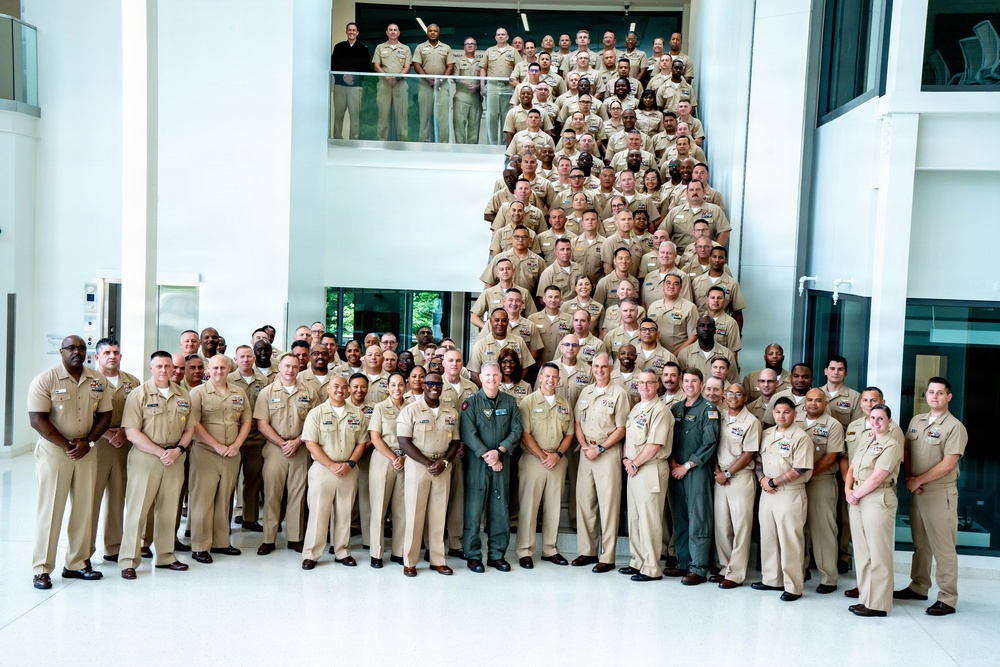 A large group photograph of what appears to be U.S. Navy personnel in khaki service uniforms, posed on and around a grand staircase inside a modern building. The majority wear standard Navy Service Uniform (NSU) khaki, with one individual in the front center wearing a green flight suit. The setting appears to be an official military facility with a bright, contemporary interior.