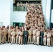 A large group photograph of what appears to be U.S. Navy personnel in khaki service uniforms, posed on and around a grand staircase inside a modern building. The majority wear standard Navy Service Uniform (NSU) khaki, with one individual in the front center wearing a green flight suit. The setting appears to be an official military facility with a bright, contemporary interior.