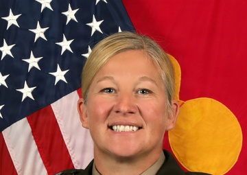 This is a formal U.S. Army official portrait of an officer identified by nameplate as 'DeLong'. She is wearing the Army Service Uniform (ASU) in green with multiple decorations, badges, and ribbons visible. The background features the U.S. flag and what appears to be a unit or general officer flag.