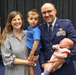 A U.S. Air Force officer in dress blues poses with his family during what appears to be a military ceremony or promotion event. He holds a newborn infant while a young toddler boy stands between him and a smiling woman, presumably his spouse. An American flag is visible in the background, suggesting an official military setting.