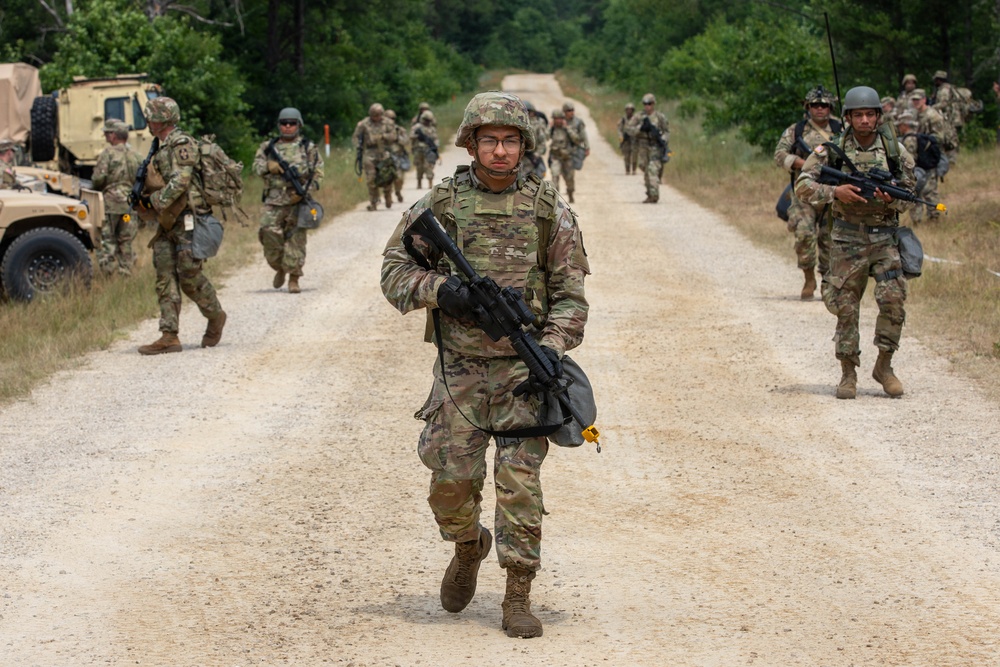 A large group of soldiers in U.S. Army OCP (Operational Camouflage Pattern) uniforms are conducting a road march along a sandy dirt trail flanked by trees and brush. The soldiers are armed and equipped with tactical gear, helmets, and body armor. A military HMMWV (Humvee) vehicle is visible on the left side of the formation.