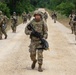 A large group of soldiers in U.S. Army OCP (Operational Camouflage Pattern) uniforms are conducting a road march along a sandy dirt trail flanked by trees and brush. The soldiers are armed and equipped with tactical gear, helmets, and body armor. A military HMMWV (Humvee) vehicle is visible on the left side of the formation.