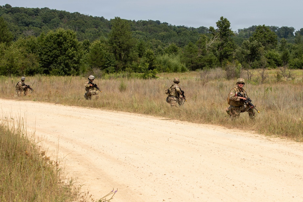 Four soldiers in camouflage uniforms are conducting a patrol along a dirt road in a rural, wooded training area. They are spaced apart in a tactical formation along the edge of the road, each carrying rifles and wearing full combat gear. The setting and formation suggest a military field training exercise rather than active combat.