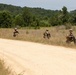 Four soldiers in camouflage uniforms are conducting a patrol along a dirt road in a rural, wooded training area. They are spaced apart in a tactical formation along the edge of the road, each carrying rifles and wearing full combat gear. The setting and formation suggest a military field training exercise rather than active combat.